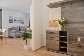 Un salon moderne avec un canapé gris, une table à manger avec des chaises, une plante en pot, un meuble en bois et un coin cuisine visible à travers une porte ouverte.