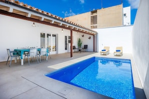 A swimming pool with a pergola, a dining area with chairs and a table, and a view of a building in the background.