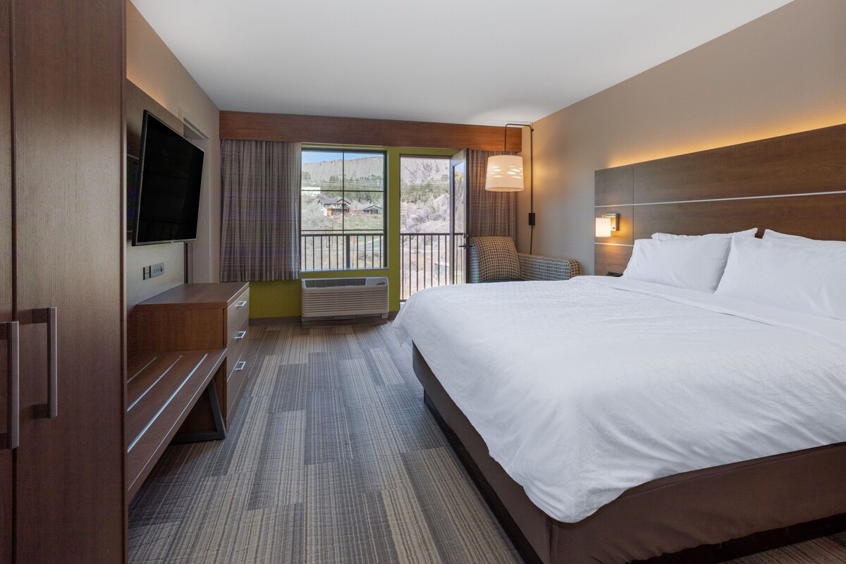 A hotel room with a large bed, a flat-screen TV, a seating area with a chair, and a balcony with a view.