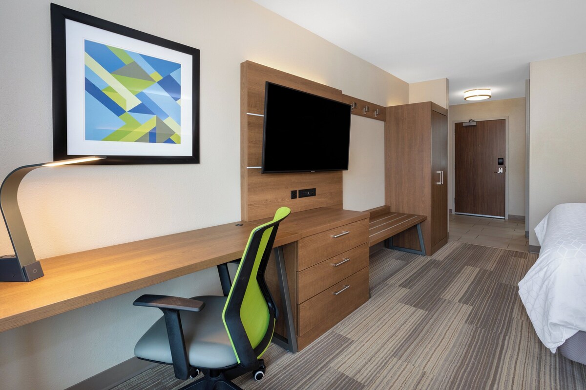 A hotel room with a wooden desk, a flat-screen TV, a modern chair, a bed, and a framed abstract art piece on the wall.