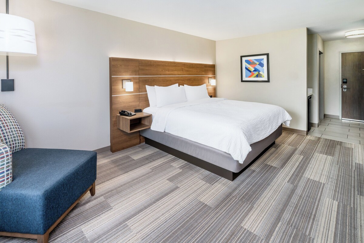 A modern hotel room with a large bed, a bedside table with a lamp, a chair, and a wooden headboard.