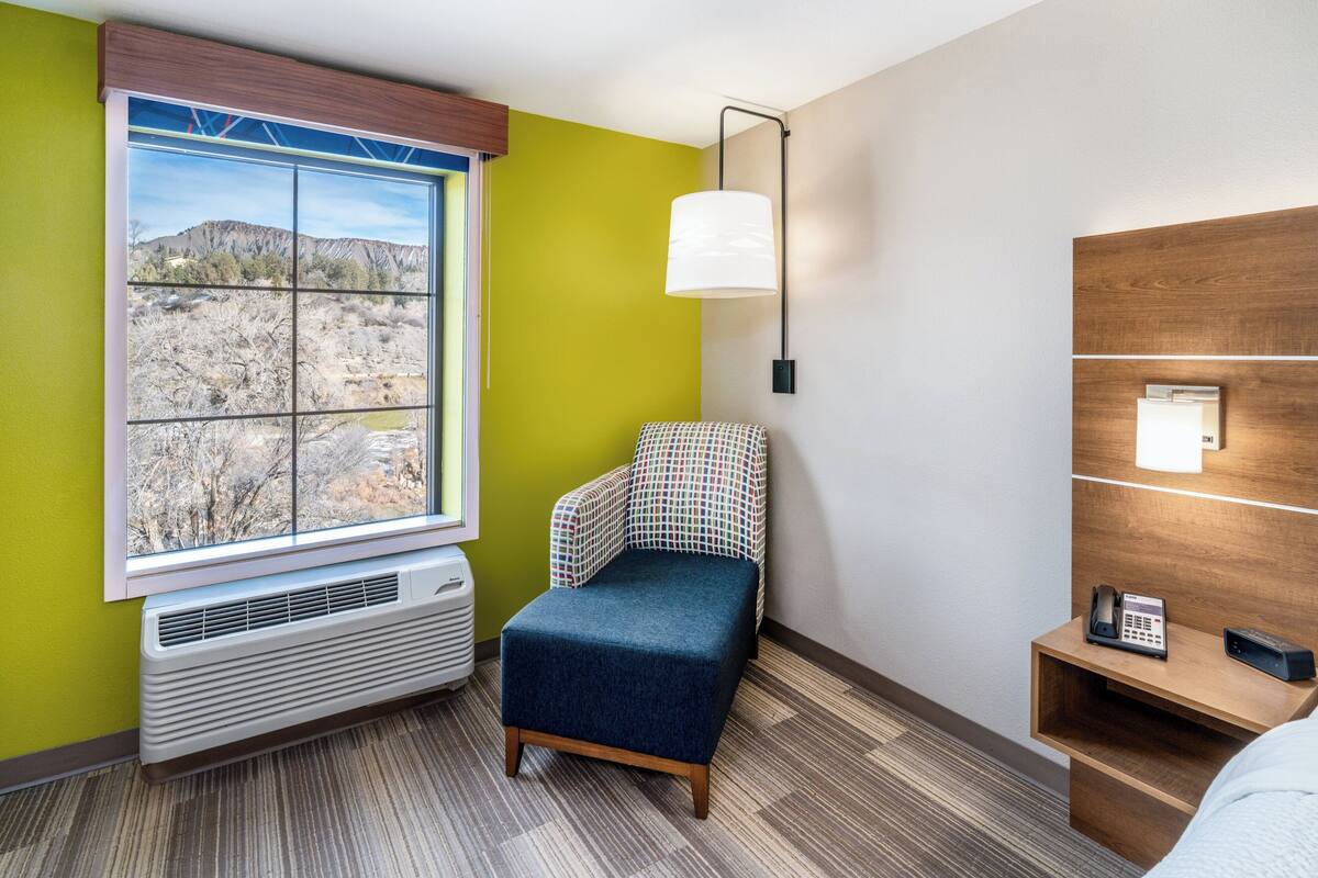 A hotel room with a large window, a chair, a bedside table with a phone, and a wall-mounted lamp.