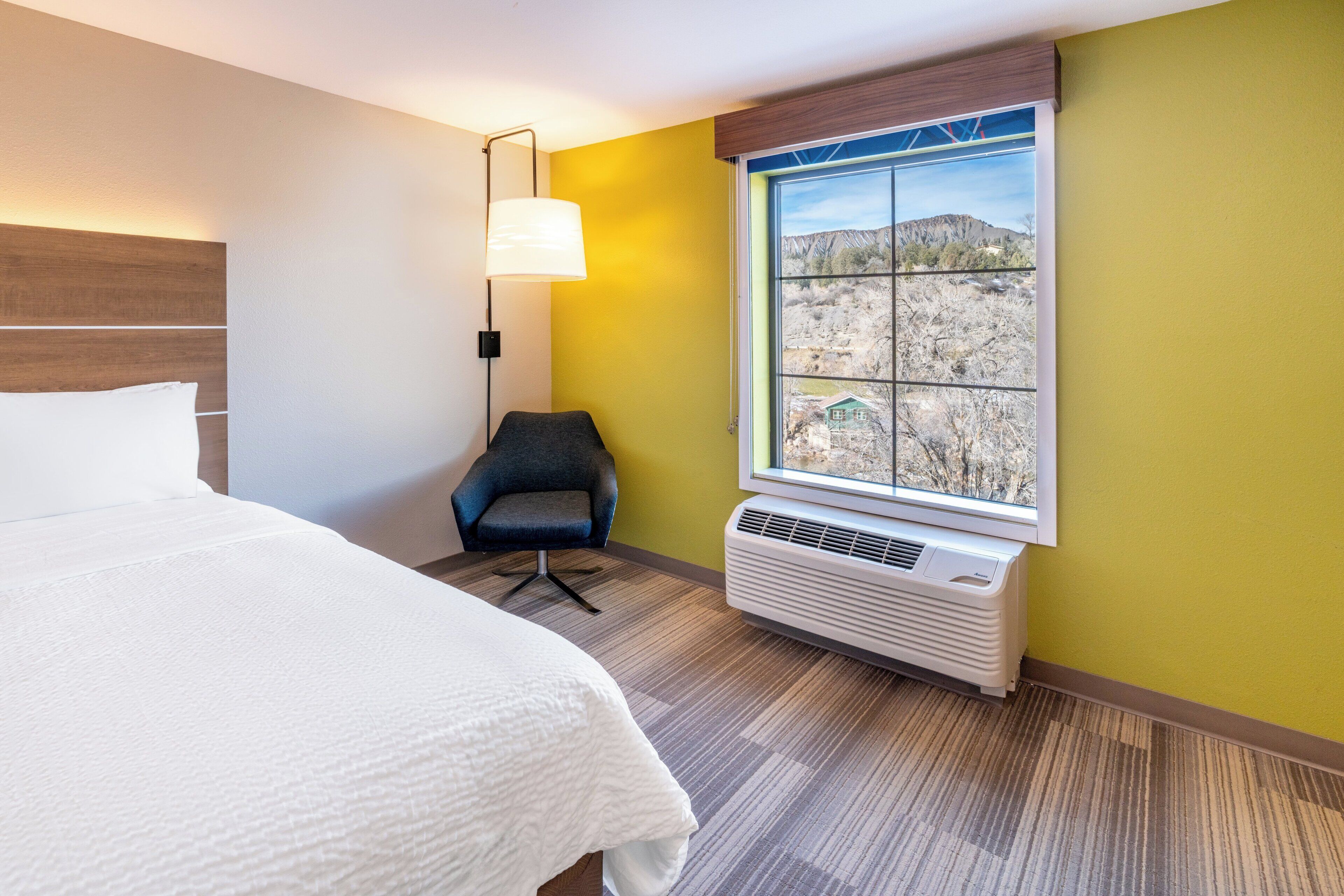 Habitación de hotel con una cama, una silla, un ventanal con vistas a un paisaje montañoso y un aire acondicionado de pared.