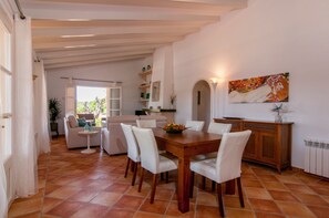 A dining area with a wooden table and chairs, a kitchen in the background, and a painting on the wall.