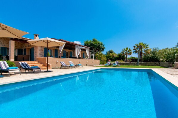 Une piscine avec des chaises longues, un patio couvert et un bâtiment en pierre, le tout sous un ciel d’un bleu limpide.