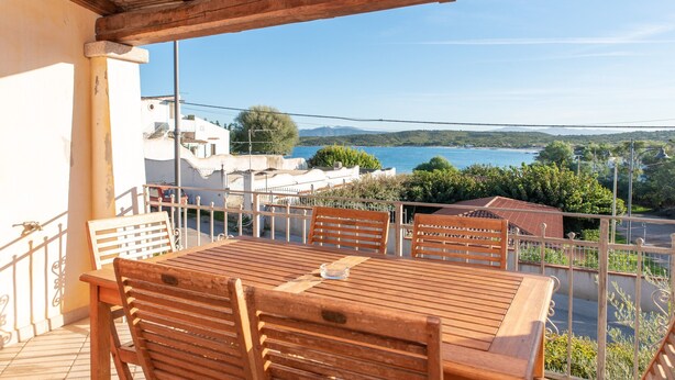 Ein Holztisch mit Stühlen auf einem Balkon, mit Blick auf ein Gewässer und Gebäude.