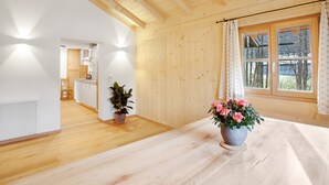 Una habitación amplia con paredes y techo de madera, un ventanal con cortinas, una mesa de madera con una planta en maceta, y vista a una cocina al fondo.