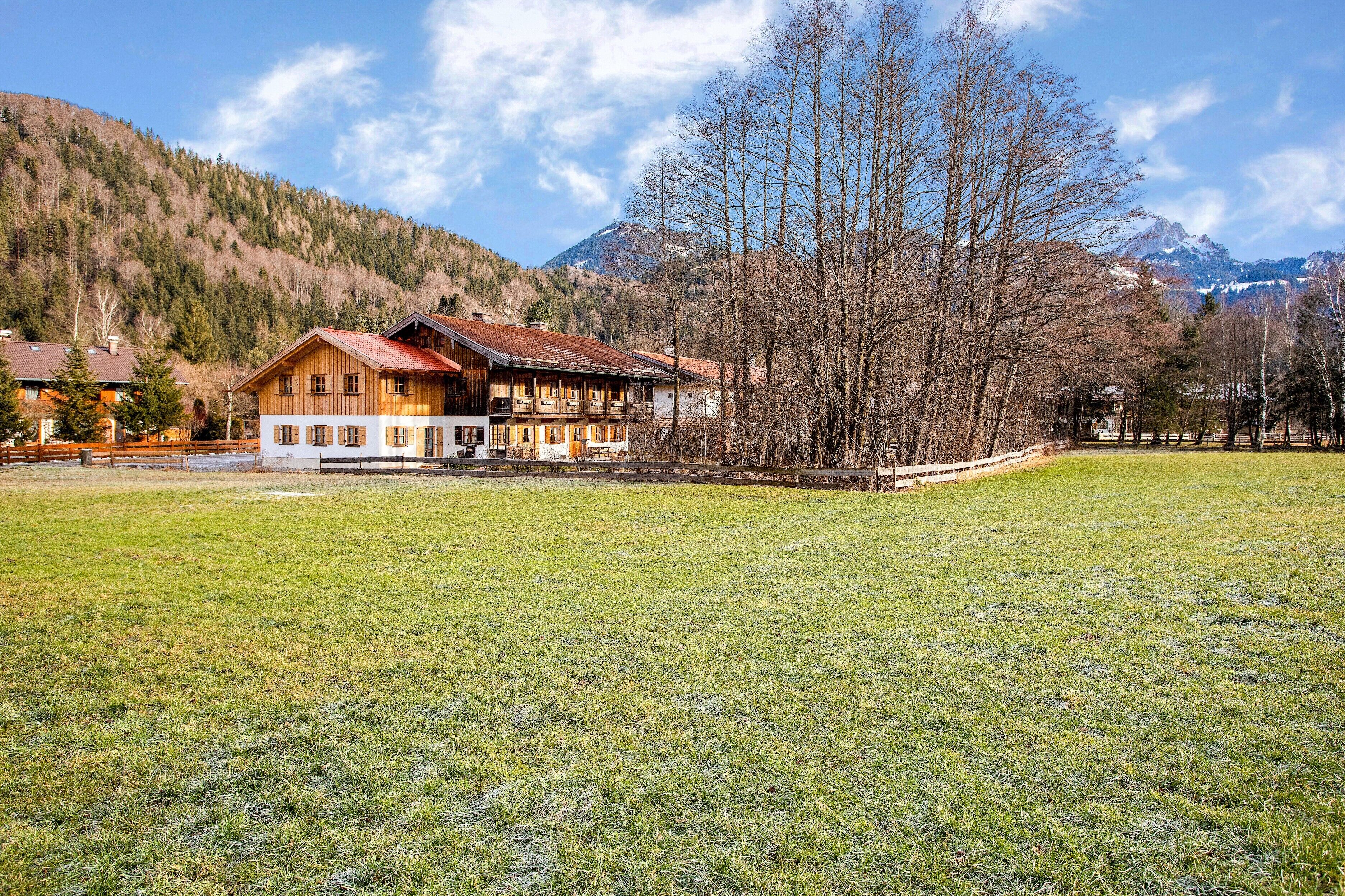 A wooden house with a balcony, surrounded by a grassy field and trees, with mountains in the background.