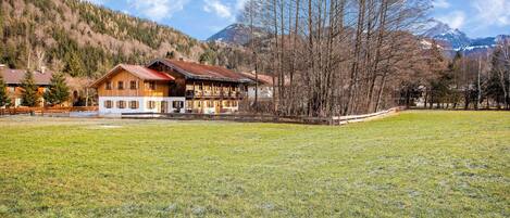 A wooden house with a balcony, surrounded by a grassy field and trees, with mountains in the background.