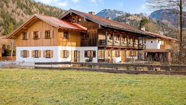 Un chalet de madera grande con varios balcones y una sección inferior blanca, con montañas al fondo.