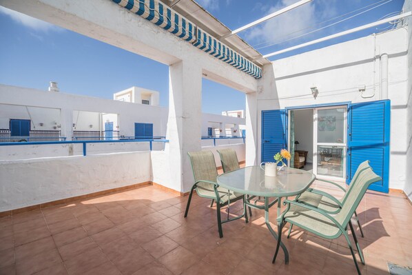 Una terraza con una mesa y sillas, una puerta azul y un edificio blanco con contraventanas azules.