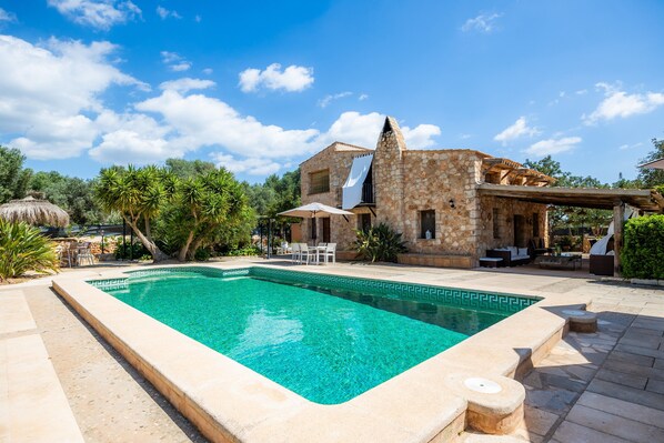 Una villa de piedra con piscina, mobiliario exterior y una estructura con techo de paja.