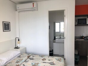 A bedroom with a bed, a bedside lamp, a white wall, and an air conditioner.