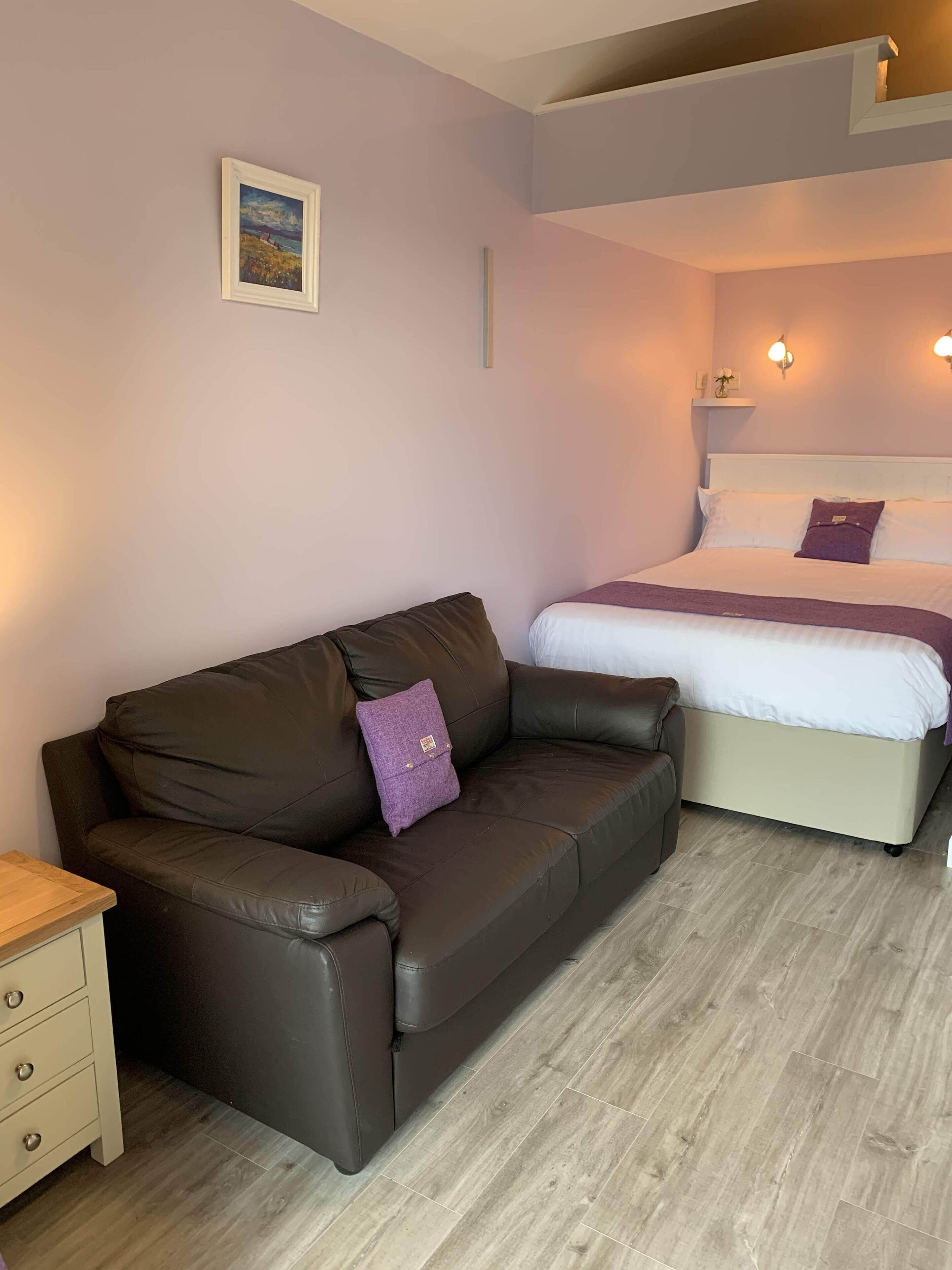 A room with a brown leather sofa, a bed with white bedding, a small wooden nightstand, and a wall-mounted shelf with a vase.
