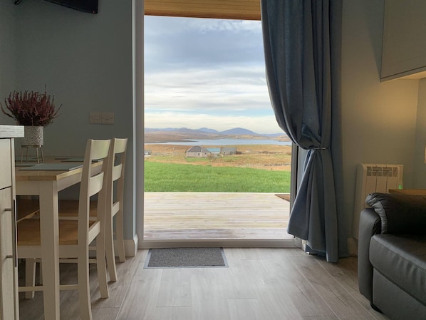 A room with a dining table and chairs, a sofa, a view of a green field and mountains through a large window, and a radiator. - Hebridean Holiday Cabins (Isle of Lewis)