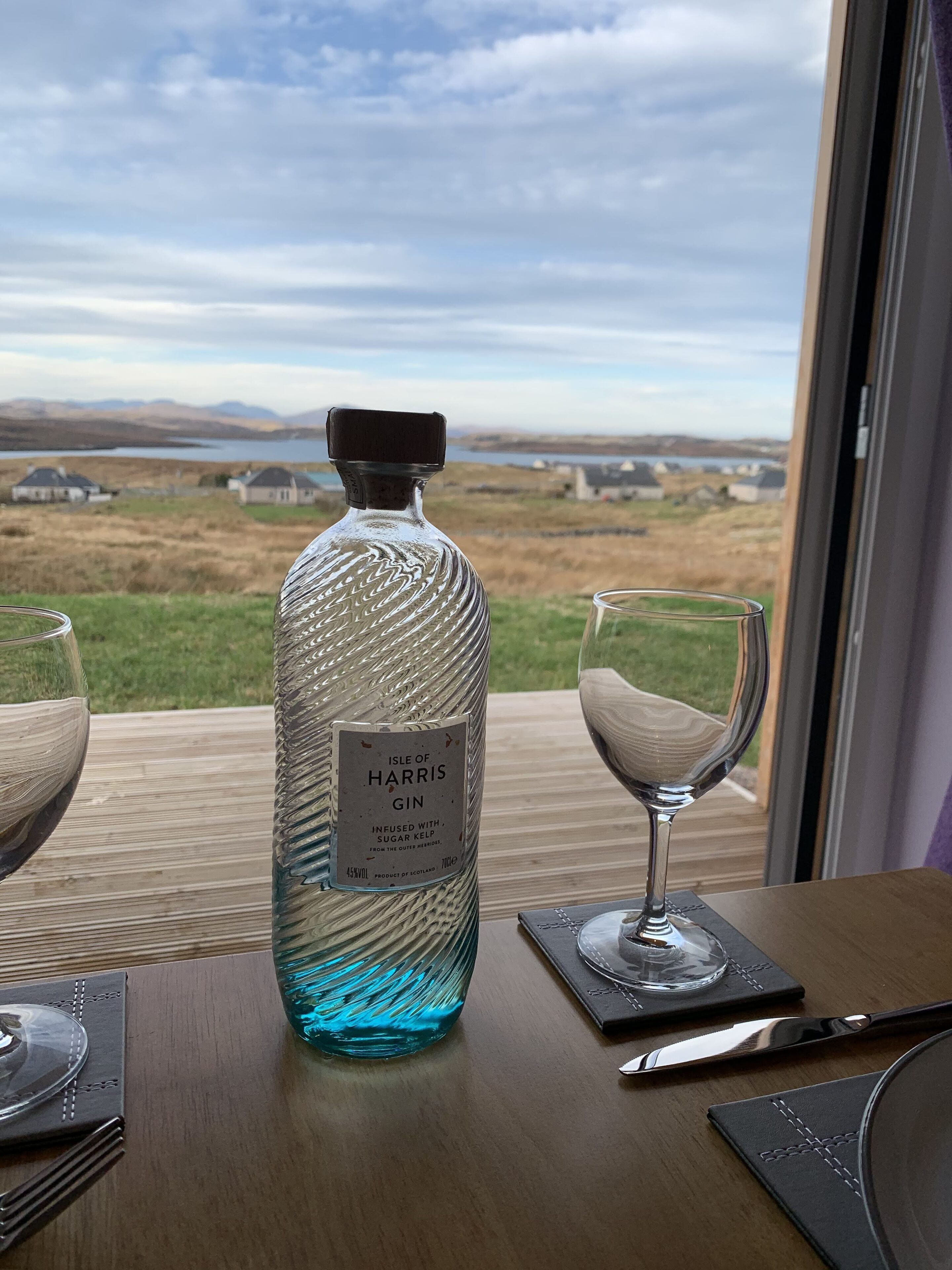 Una bottiglia di gin Isle of Harris su un tavolo di legno, con vista su un paesaggio attraverso una finestra.