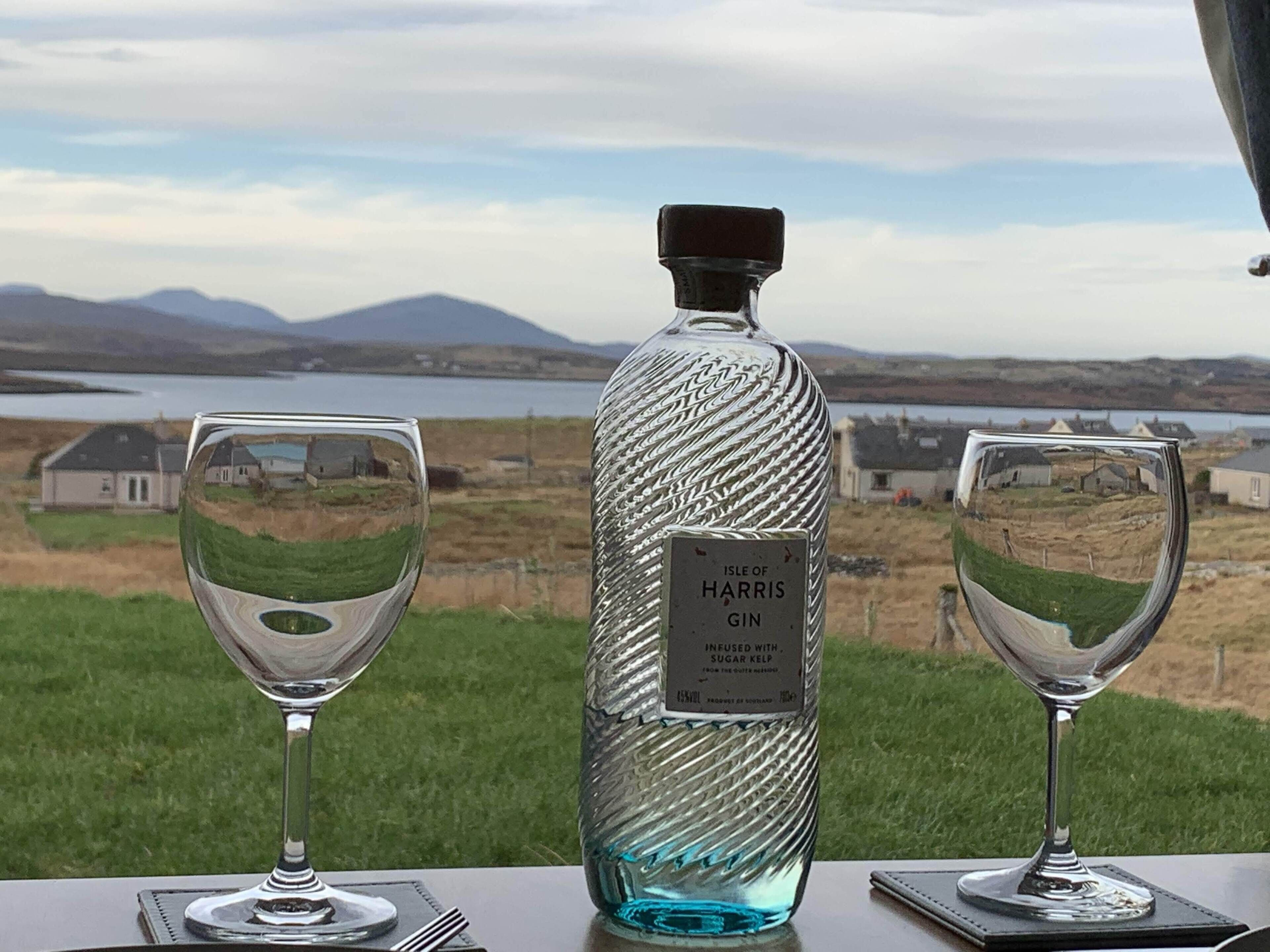 A bottle of Harris Gin and two glasses on a table with a scenic view of houses and mountains in the background.