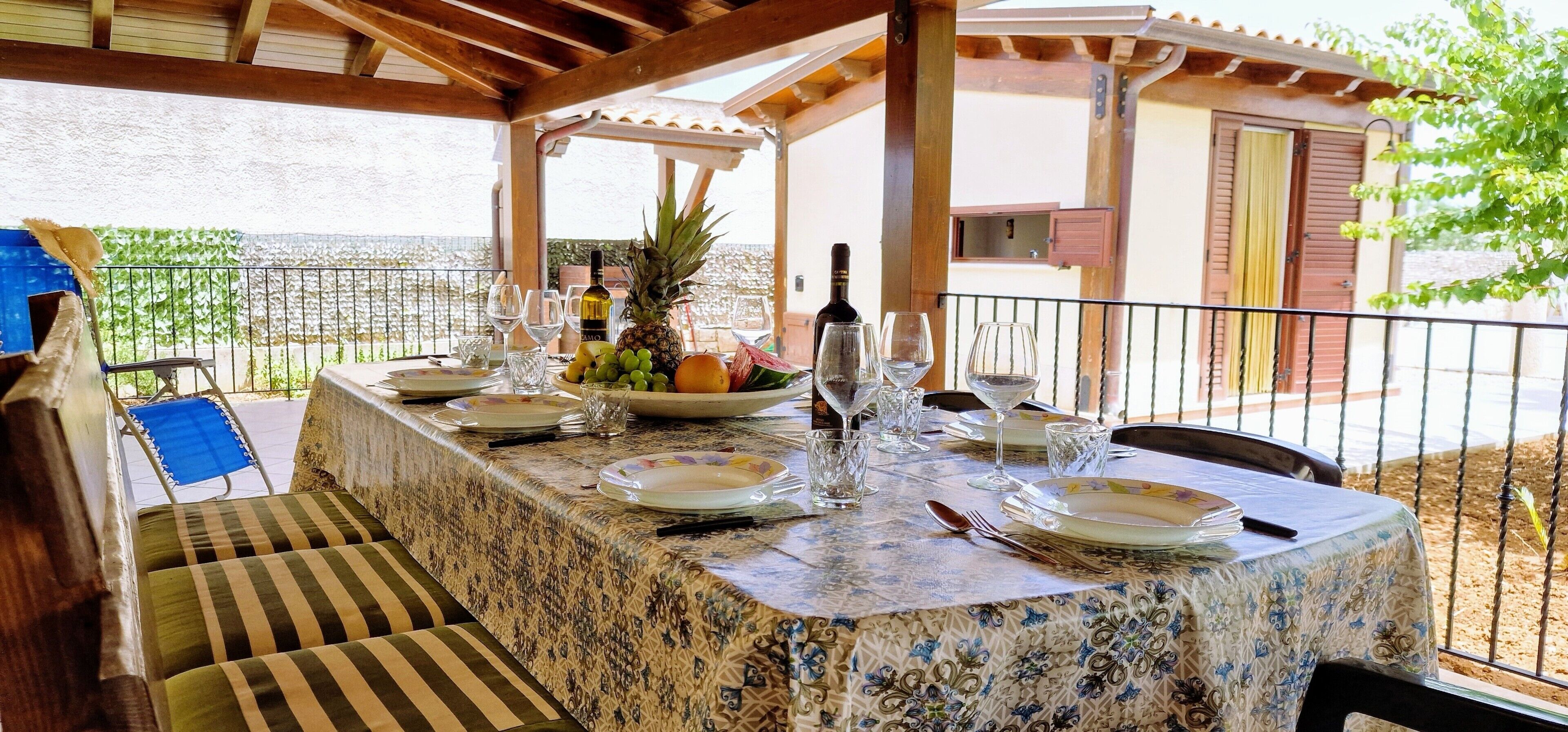 Un espace extérieur abrité, aménagé pour les repas, avec une table dressée pour quatre personnes, un ananas et une bouteille de vin.