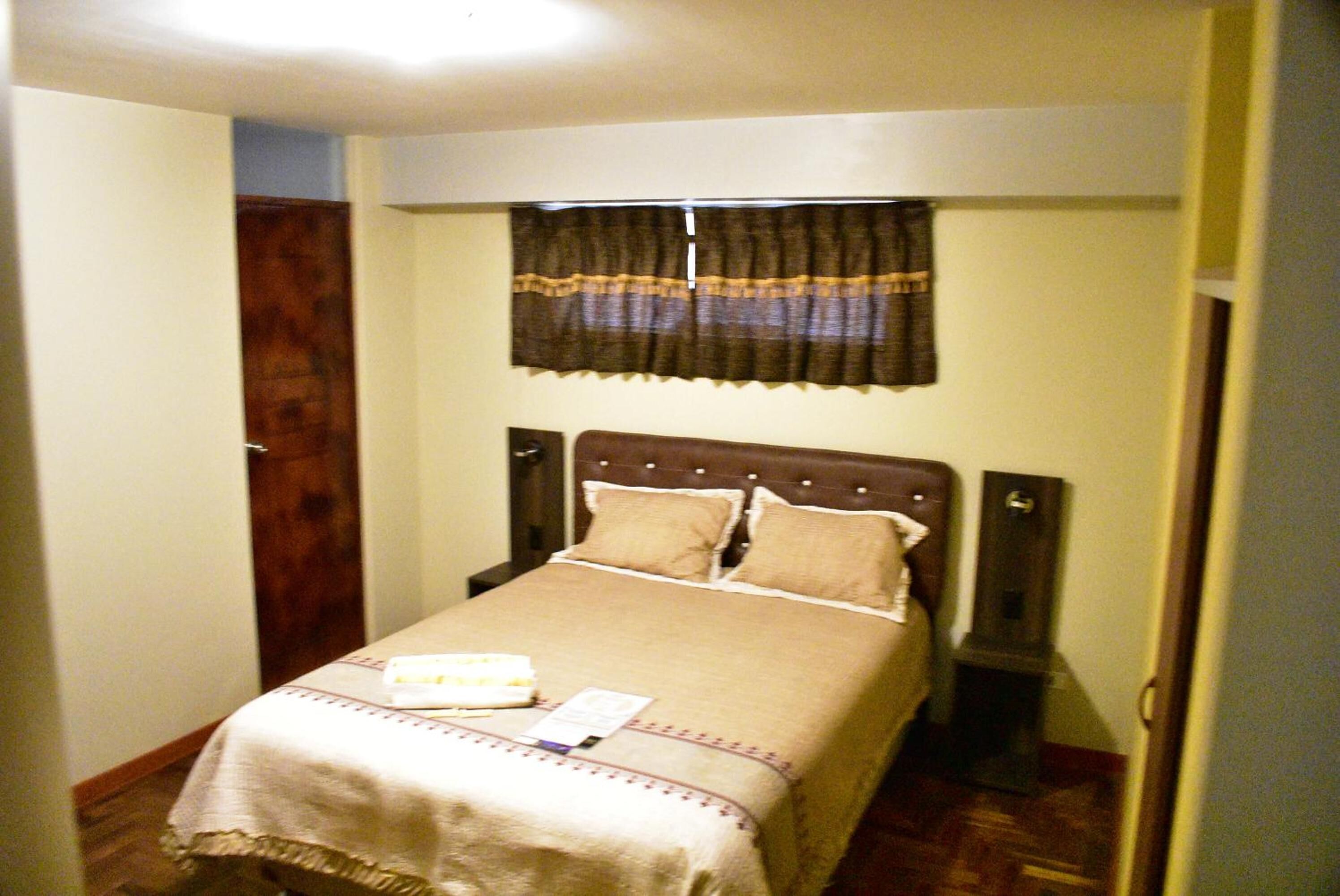 Una cama pequeña y ordenada con cabecera marrón y ropa de cama color beige. Se ve una ventana con cortina oscura sobre la cama.