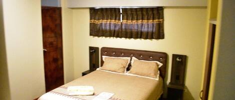 Una cama pequeña y ordenada con cabecera marrón y ropa de cama color beige. Se ve una ventana con cortina oscura sobre la cama.