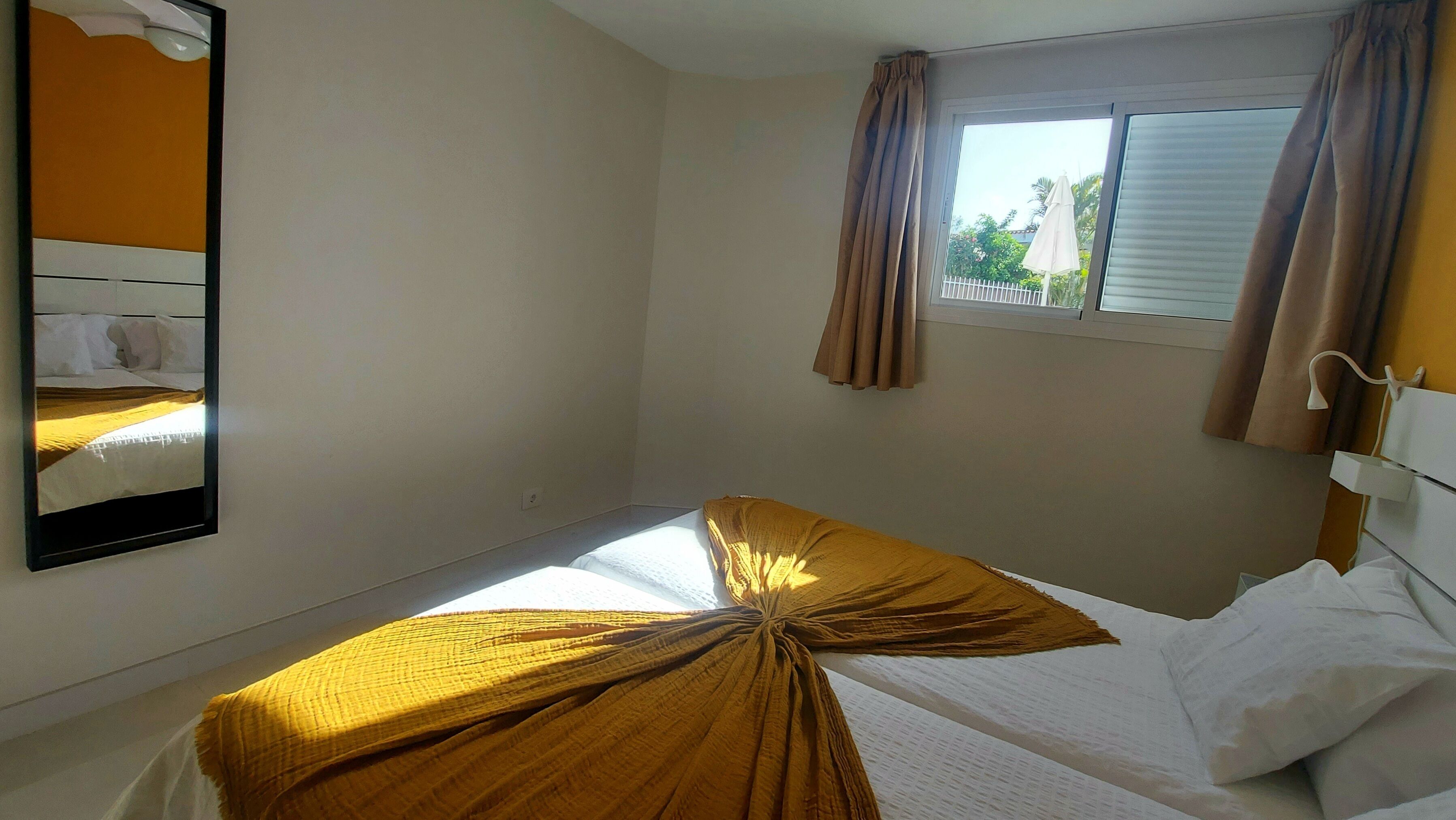 A bedroom with a single bed, a window with curtains, a mirror on the wall, and a small table.