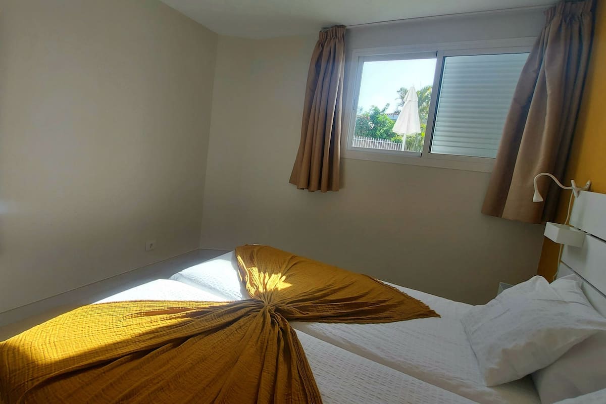A bedroom with a single bed, a window with curtains, a mirror on the wall, and a small table.