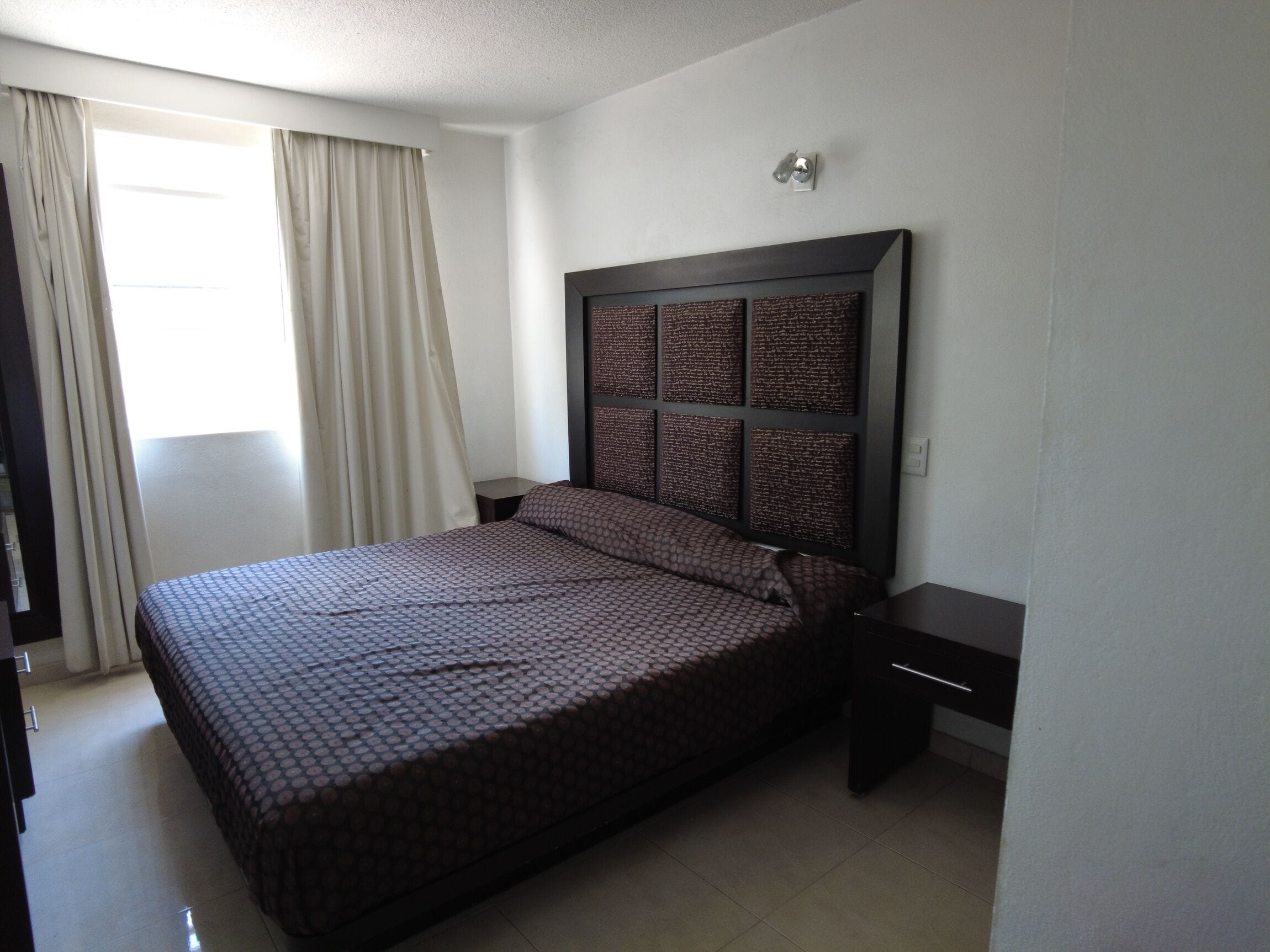 Habitación de hotel con una cama grande, una mesita de noche, una lámpara de pared y una ventana con cortinas.