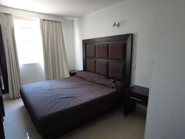 Habitación de hotel con una cama grande, una mesita de noche, una lámpara de pared y una ventana con cortinas.