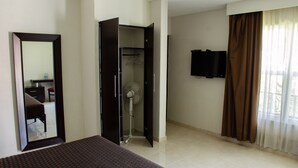 A hotel room with a bed, a wardrobe, a mirror, a fan, a television, and a window with curtains.