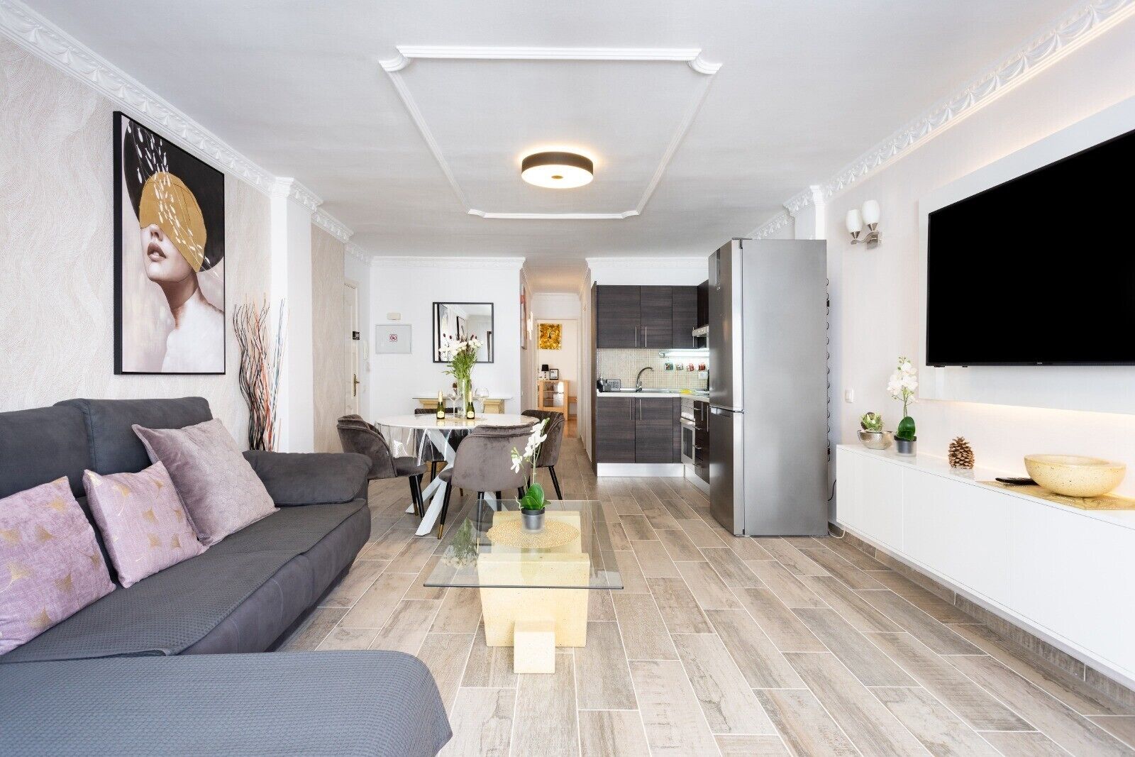 Una sala de estar moderna con un sofá gris, un comedor con una mesa redonda y una cocina con armarios oscuros y un refrigerador de acero inoxidable.