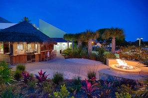 Relax in our resort's serene garden, surrounded by lush greenery and a central fire feature, near the thatched-roof bar and lounge area.