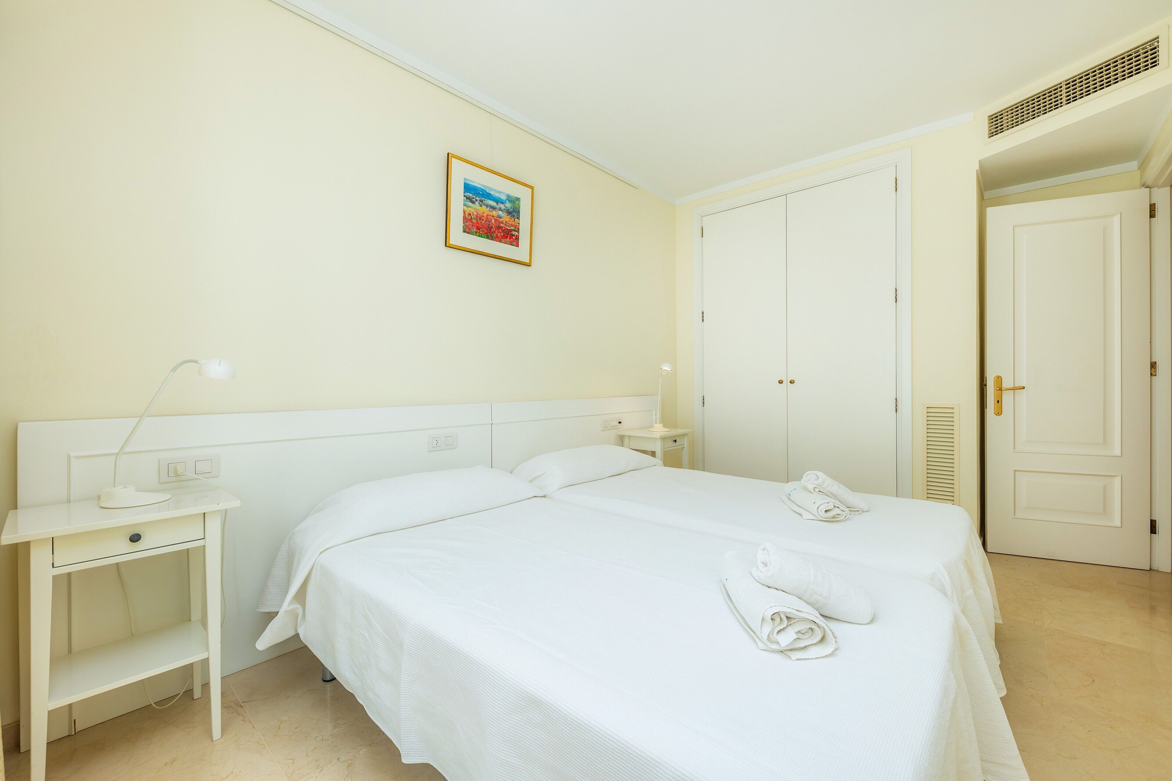 Una habitación de hotel con una cama individual, una mesita de noche con lámpara, un cuadro en la pared y una puerta hacia otra habitación.