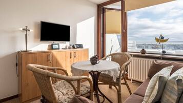 A room with a television, a table with a coffee mug, wicker chairs, a sofa, and a view of the city through a window.