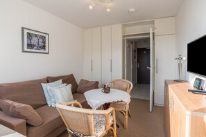 A living area with a brown sofa, wicker chairs, a small table with a white tablecloth, a framed picture on the wall, and a TV on a wooden cabinet.