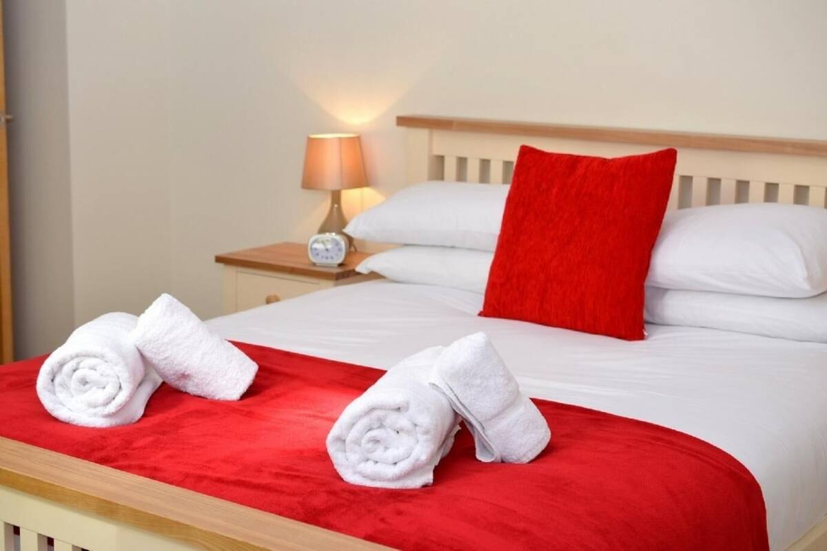 A neatly made bed with white linens and a red pillow, a bedside lamp, and two rolled towels on the bed.