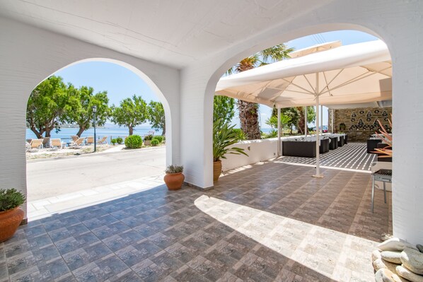 A covered outdoor area with a view of the sea, featuring a seating area with tables and chairs, potted plants, and a tiled floor.