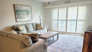 A living room with a beige sofa, a coffee table, and a rug. There is a painting on the wall and a window with blinds. - Ocean view 2 bedroom, 2.5 bath townhouse. Nicely upgraded! (St. Augustine)