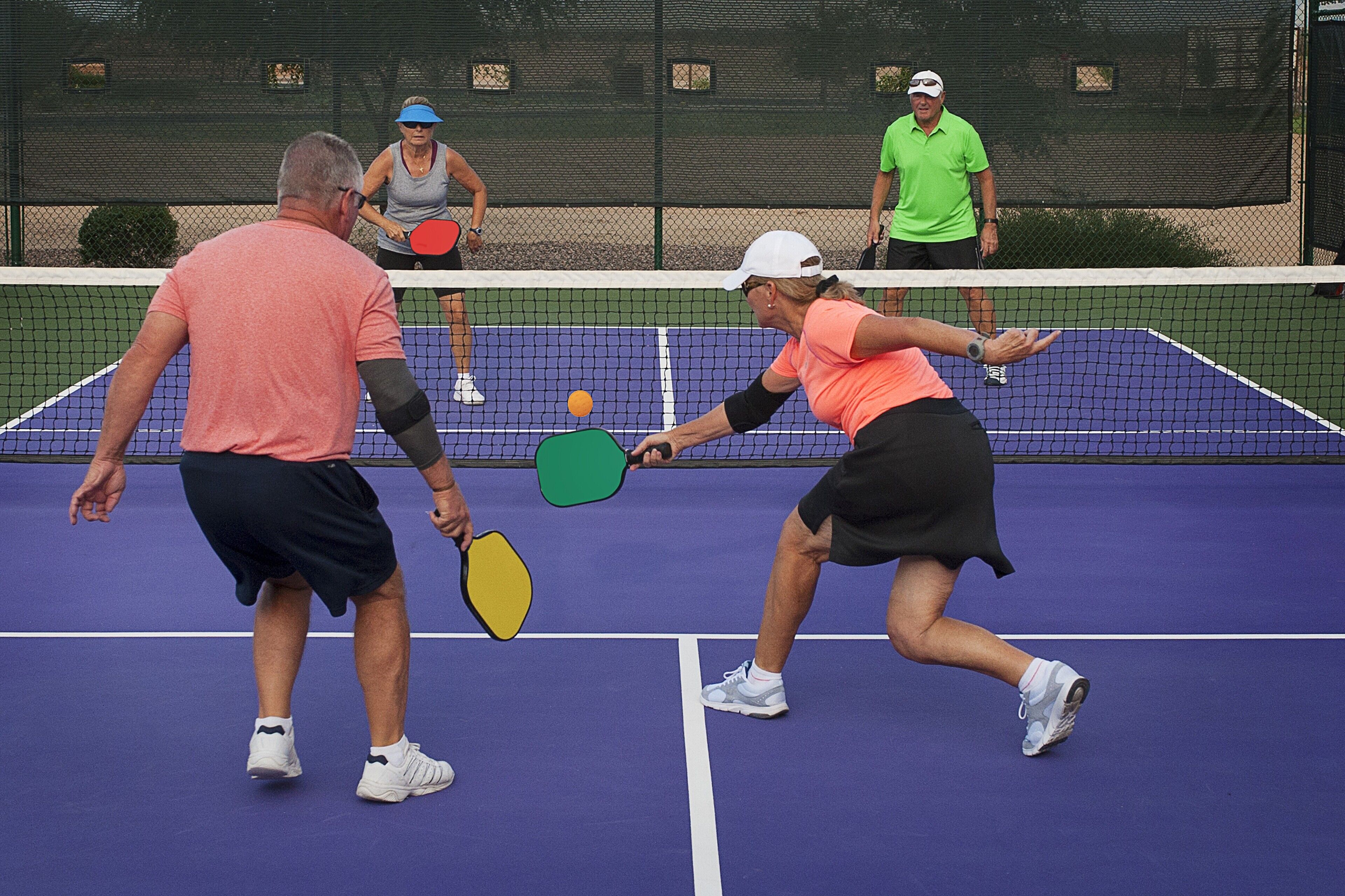 Una cancha de tenis con jugadores en movimiento, una red y una valla al fondo.
