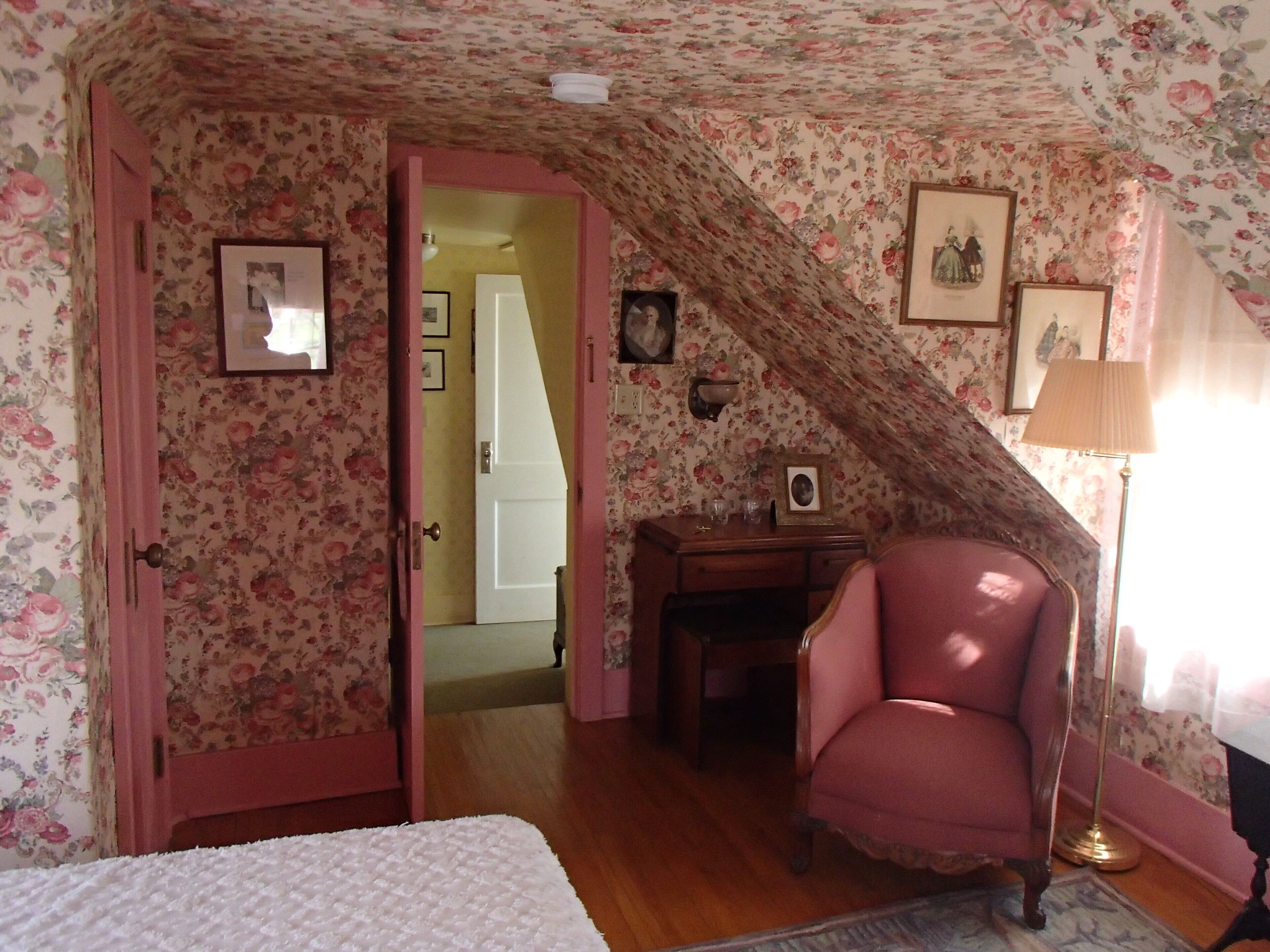 A bedroom with floral wallpaper, a pink chair, a desk, and a bed.