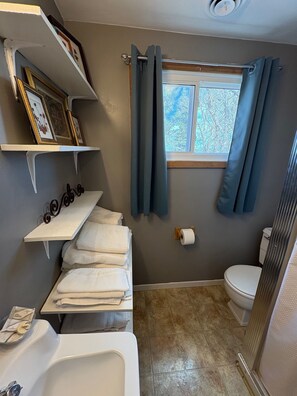 A bathroom with a bathtub, toilet, and shower area. There are towels stacked on a shelf, and a window with curtains.