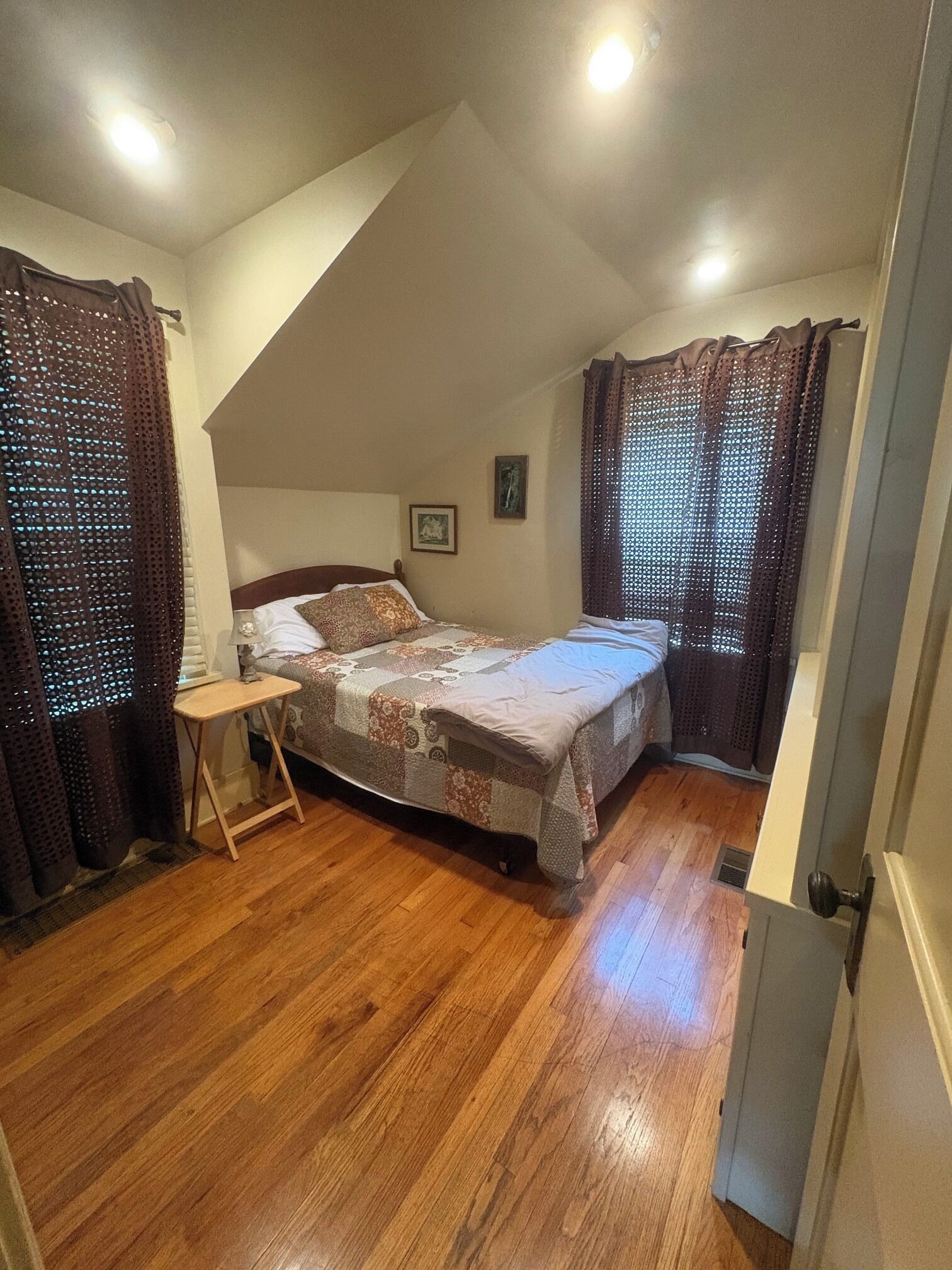 Un dormitorio con techo inclinado, una cama, una mesita de noche y un ventanal con cortinas.