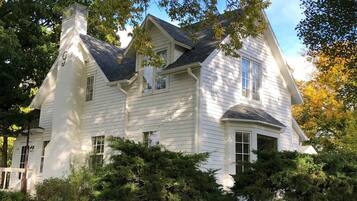 A large, white, two-story house with a steeply pitched roof, multiple windows, and surrounded by dense greenery.
