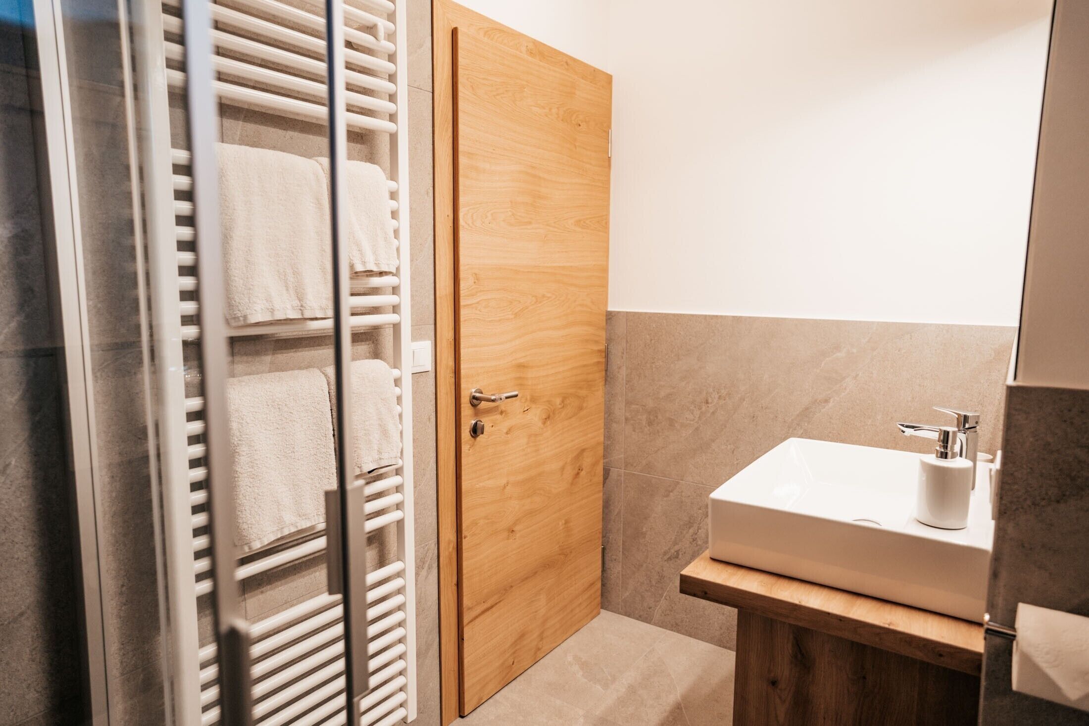 A bathroom with a wooden door, a glass shower enclosure, a white sink, and a towel rack.