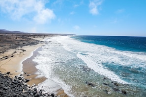 A coastal landscape with a sandy beach, rocky shoreline, and a clear blue sky.