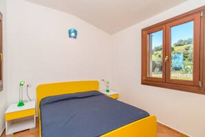 Un dormitorio con cama, mesita de noche, lámpara, ventana con vistas a la vegetación y un reloj de pared.