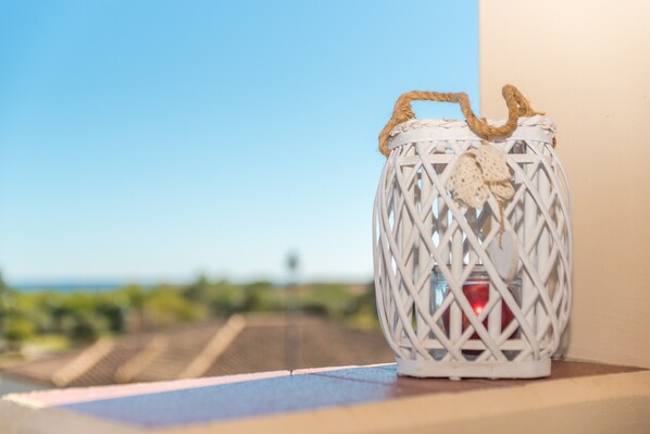 A white wicker lantern with a rope handle and a decorative element, placed on a ledge with a clear view of the ocean and greenery in the background.