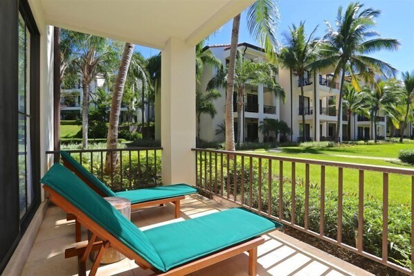 A balcony with two wooden lounge chairs and a green cushion, overlooking a well-maintained lawn and palm trees.