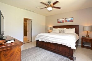 A bedroom with a wooden bed, a ceiling fan, a desk with a lamp, and a door leading to another room.