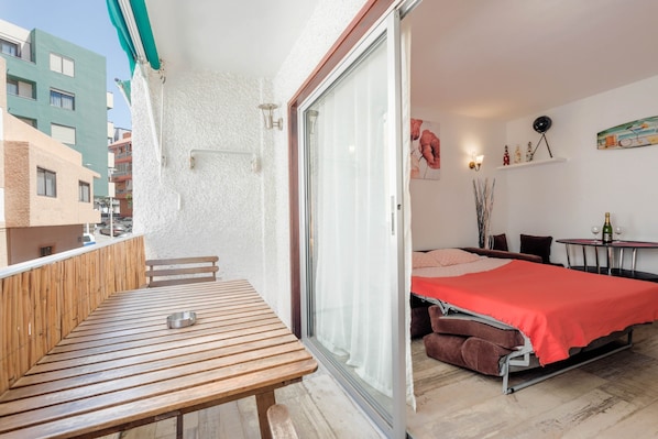Un balcón con una mesa de madera y un cuenco de metal, una puerta corrediza de vidrio que da a un dormitorio con colcha roja, y una mesita con una botella y vasos.