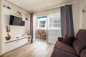 Una sala de estar con una televisión de pantalla plana, un sofá, una mesa de comedor con sillas y un ventanal corredizo que da a un área exterior.